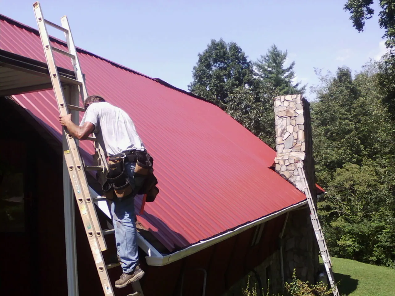 Roofer on a ladder beside a red metal roof and stone chimney — Soffit & Fascia Repair by Roofing Brothers in Jetersville